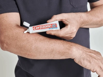 A person applies Cortizone-10 cream onto their forearm. The tube is held in one hand, cap open, ready to soothe and relieve skin irritation.